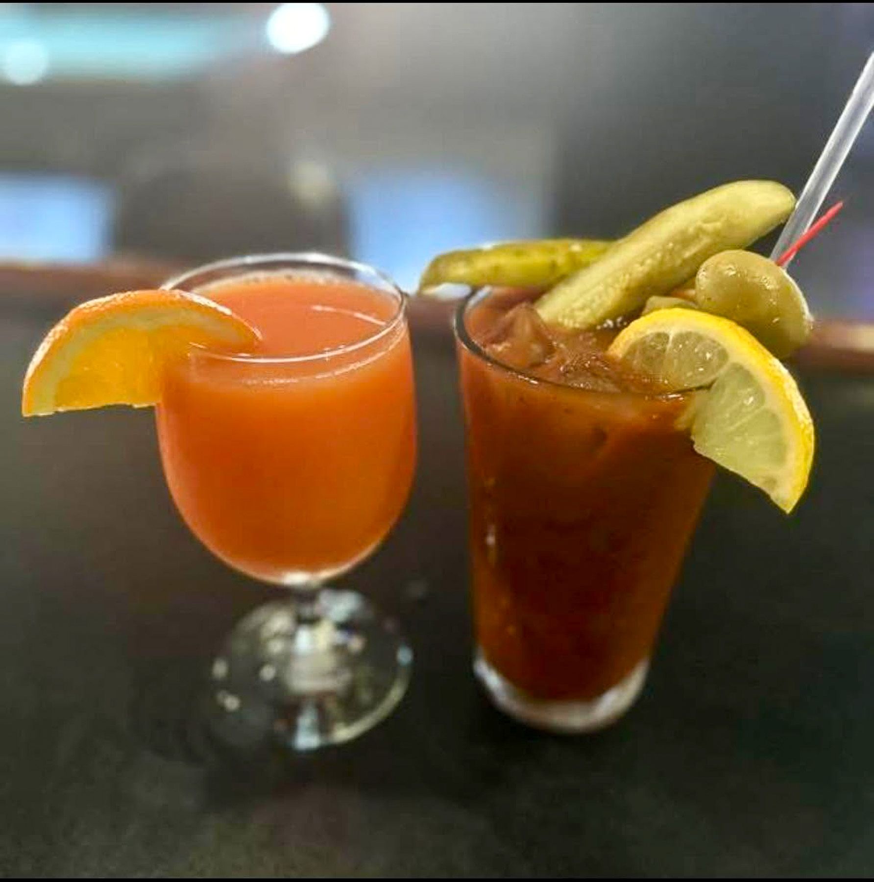 Two cocktails on a table, one orange and one dark red, both garnished with lemon wedges and pickles.