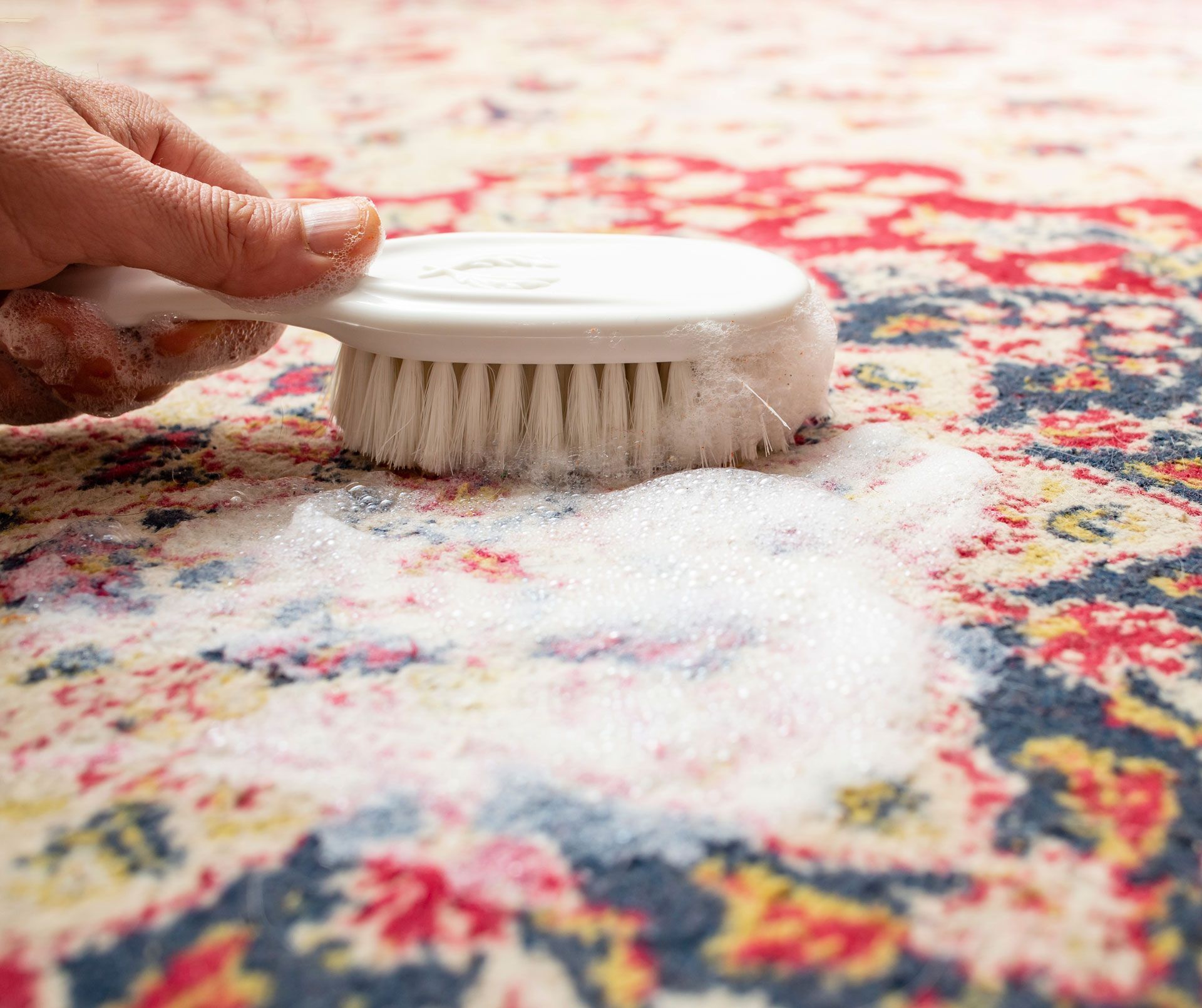 Hand scrubbing a colorful patterned rug with a white brush and soapy foam