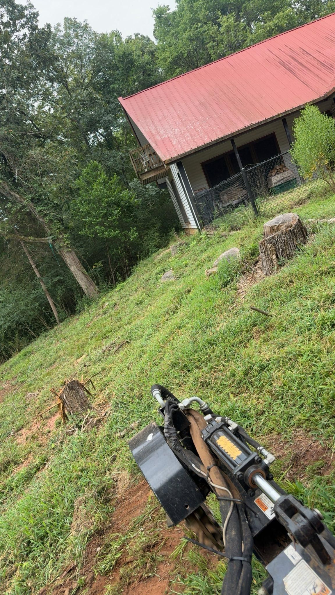 A stump grinder at work on a grassy slope with a cabin in the background.