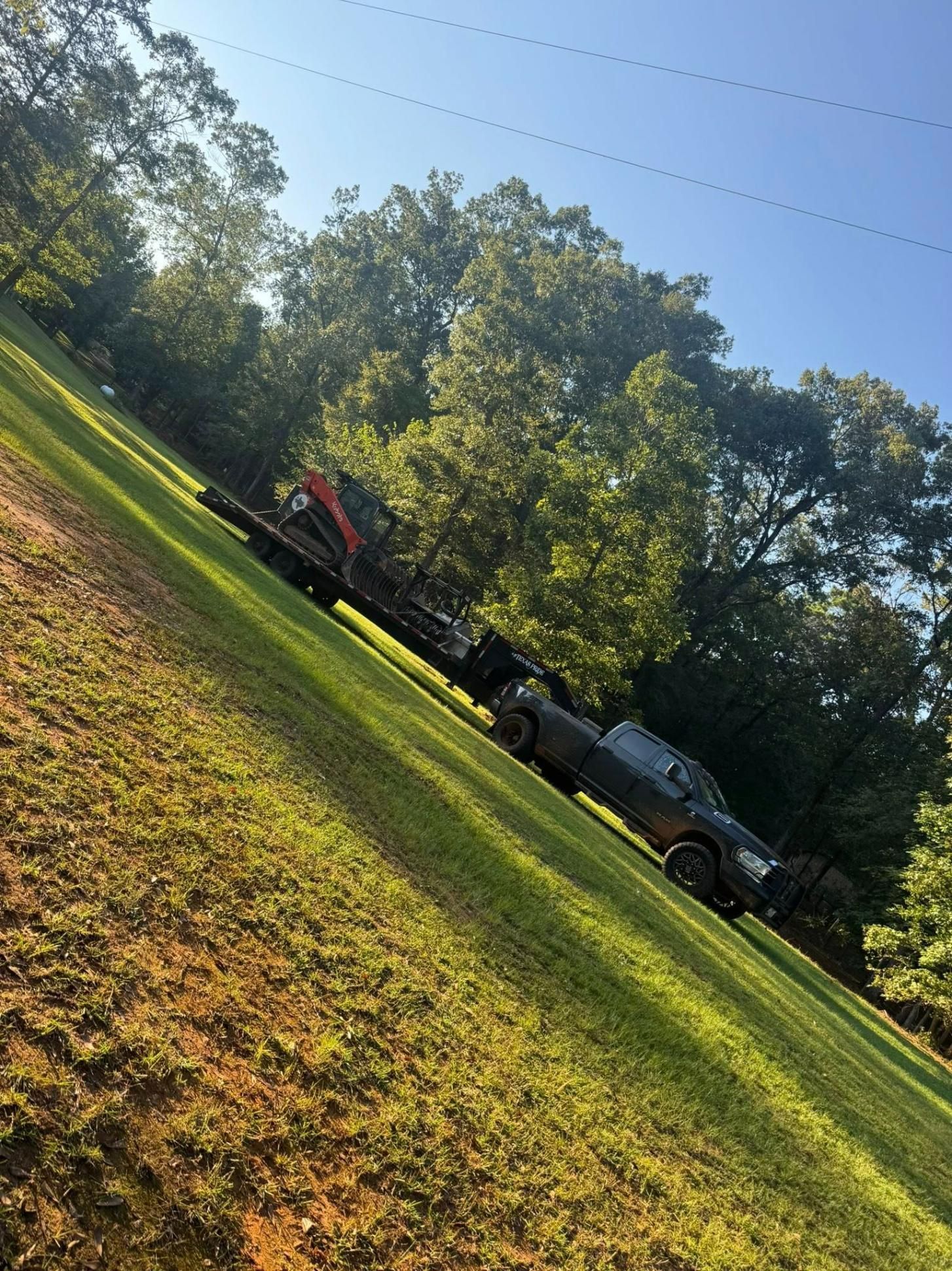 Truck pulling a trailer with equipment on a grassy hillside, trees in background, sunny day.