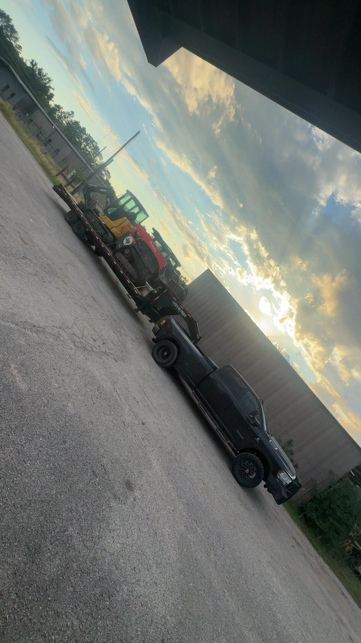 Black truck pulling a trailer with heavy machinery on it, parked in a lot, under a cloudy sky.