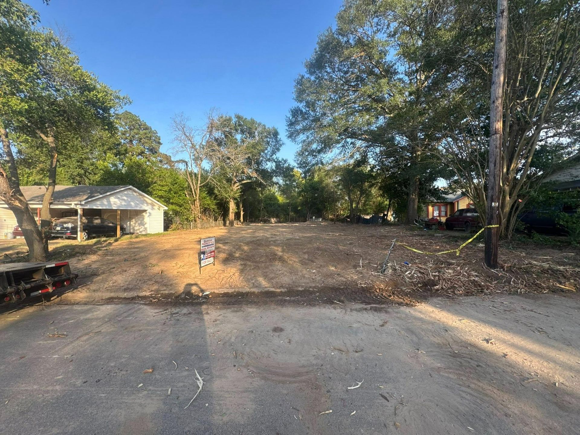 Empty lot cleared for construction, trees in background, small buildings on either side, sunny day.