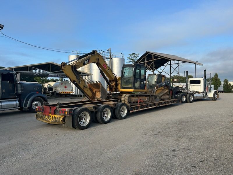 Flatbed truck hauling excavators and a metal frame in a gravel lot under a blue sky