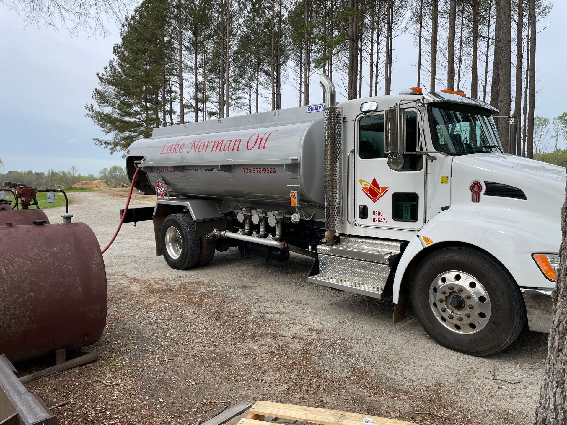 White fuel tanker truck parked on gravel near trees, with a hose connected to a large tank.