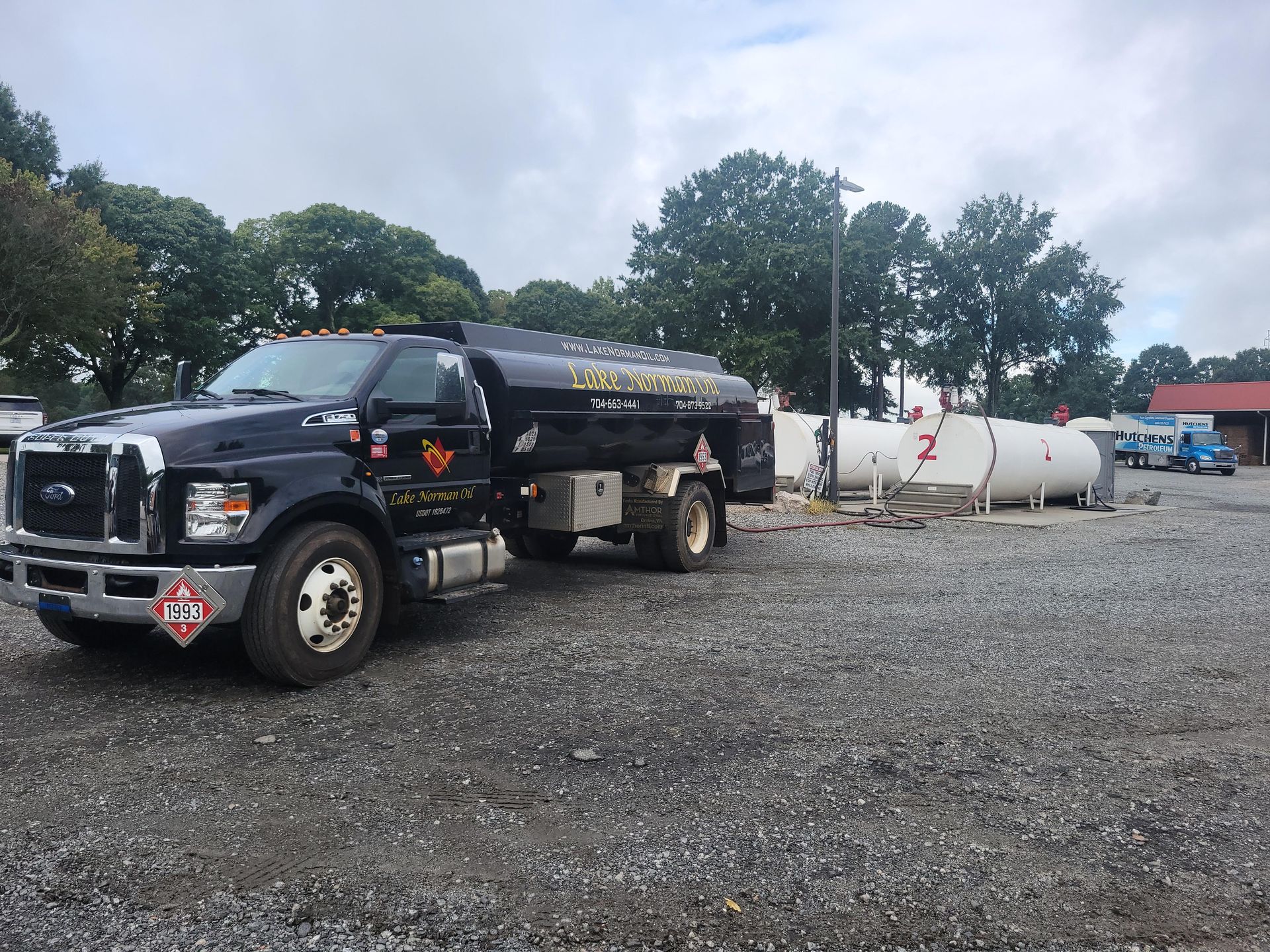 Black fuel tanker truck parked on gravel beside white storage tanks under cloudy skies