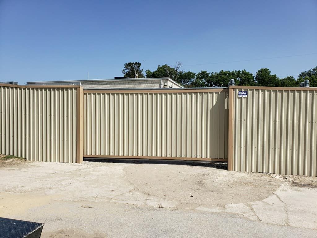 Beige corrugated metal fence with sliding gate; gravel driveway, trees, and sky in background.