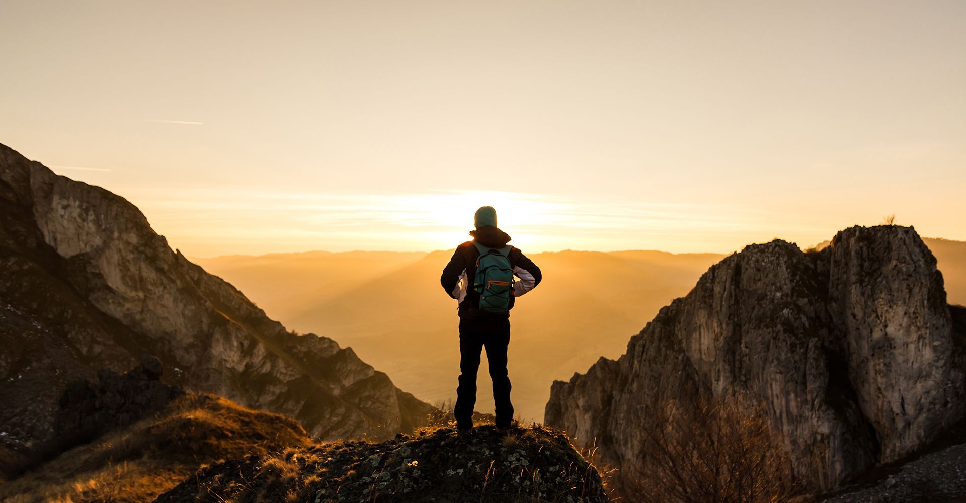 A man with a backpack is standing on top of a mountain at sunset.