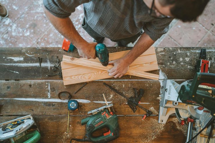 Person drilling a wooden plank at a cluttered workbench with tools and a saw nearby