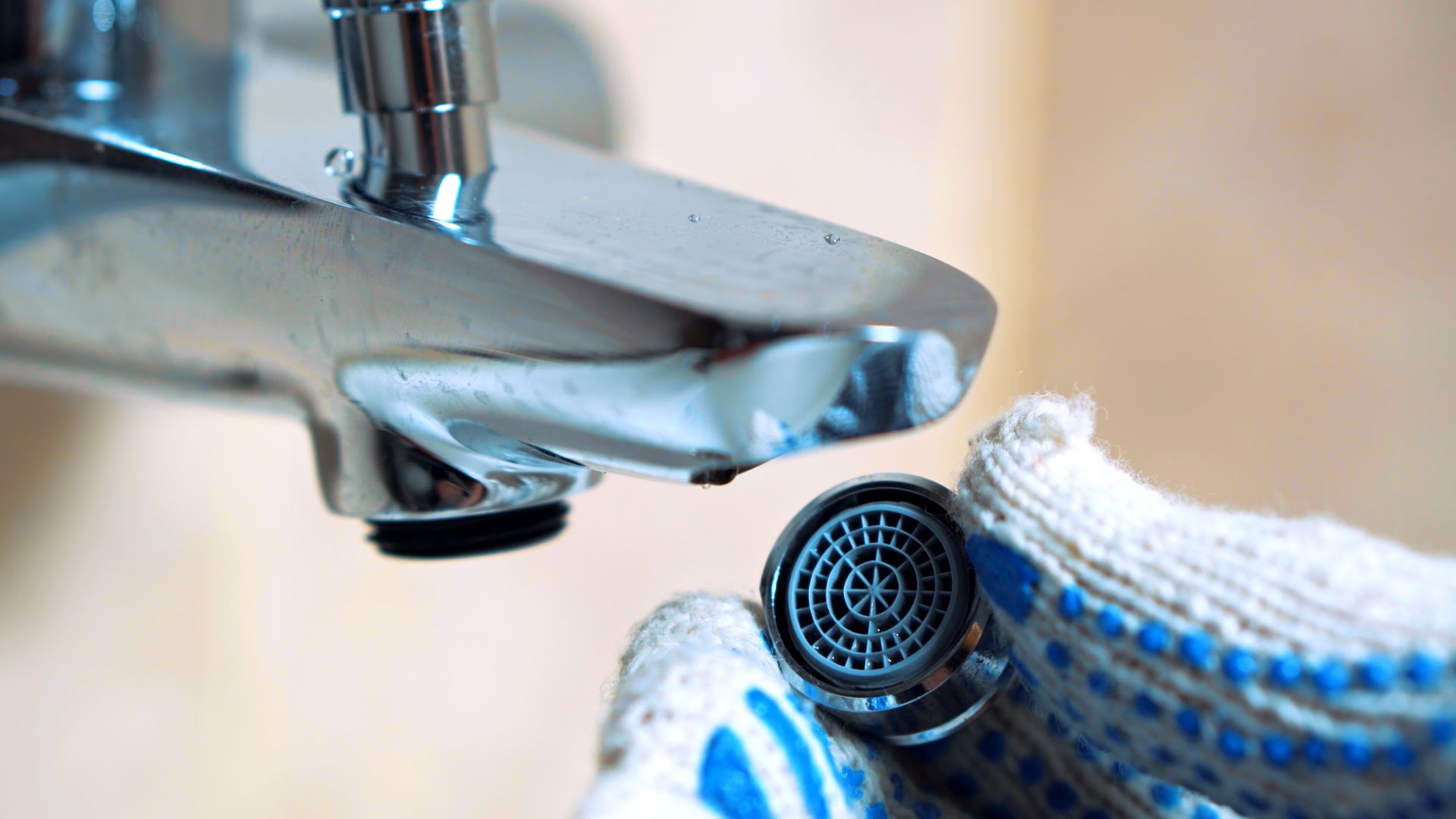 A hand in a blue glove holds a faucet aerator under a running sink tap.
