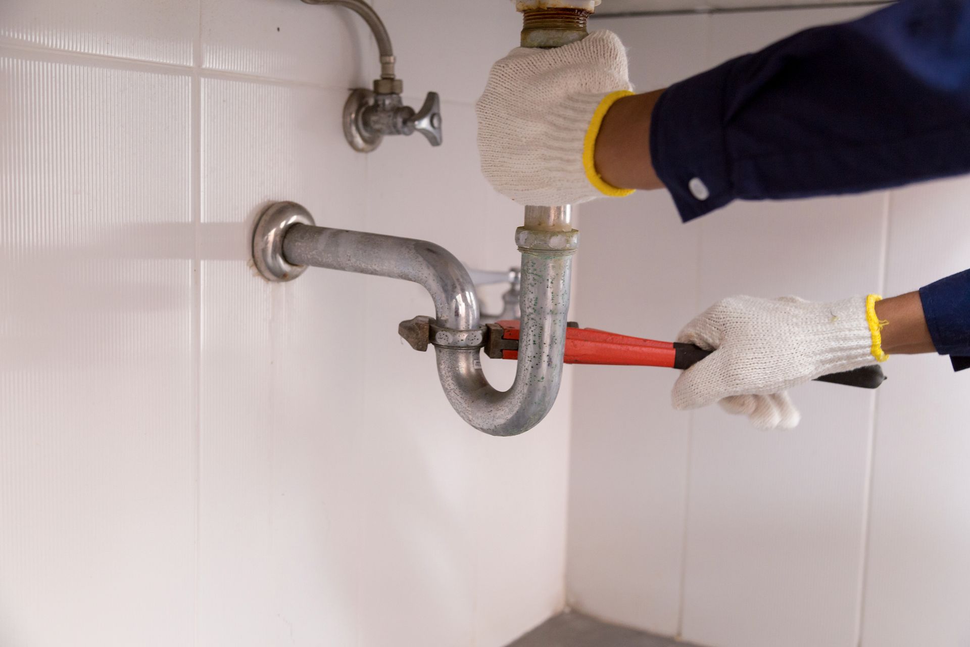 Gloved hands using a wrench to tighten a red pipe under a sink in a white tiled wall.