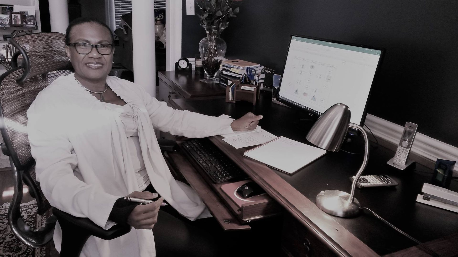 Woman in a white jacket sits at a dark desk, pointing at papers with a pen. The desk has a lamp and calendar.