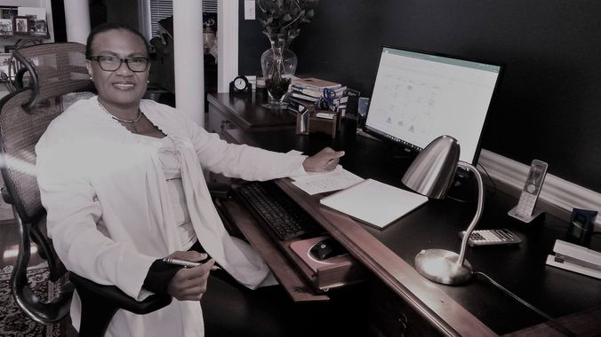 Woman in a white jacket sits at a dark desk, pointing at papers with a pen. The desk has a lamp and calendar.