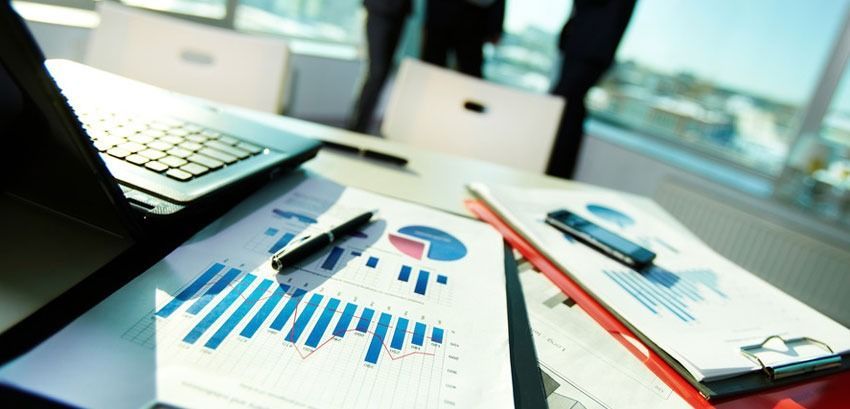 Laptop, charts, pen, and phone on a desk, with people in the background.