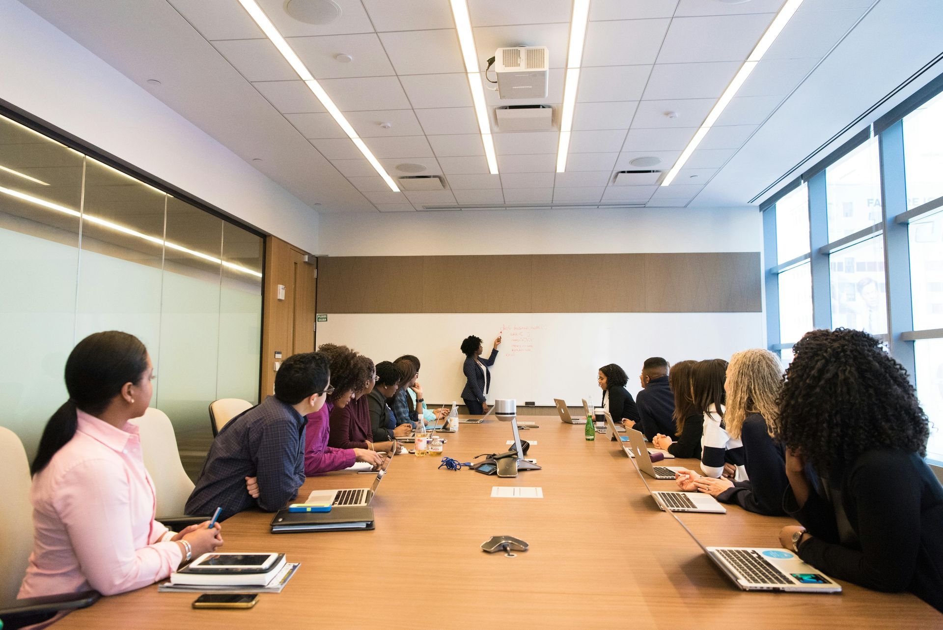 People in a boardroom, one at the whiteboard. They are listening and looking at the speaker.