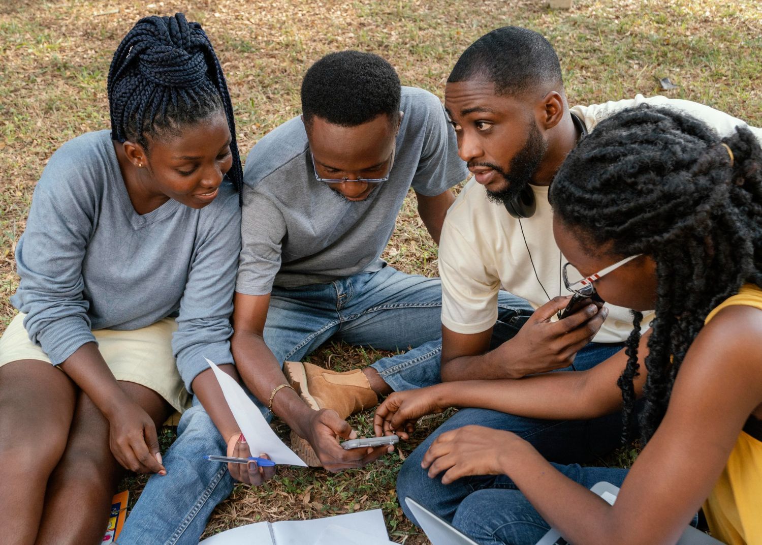 Four people sit on grass outdoors, looking at and discussing a paper together.
