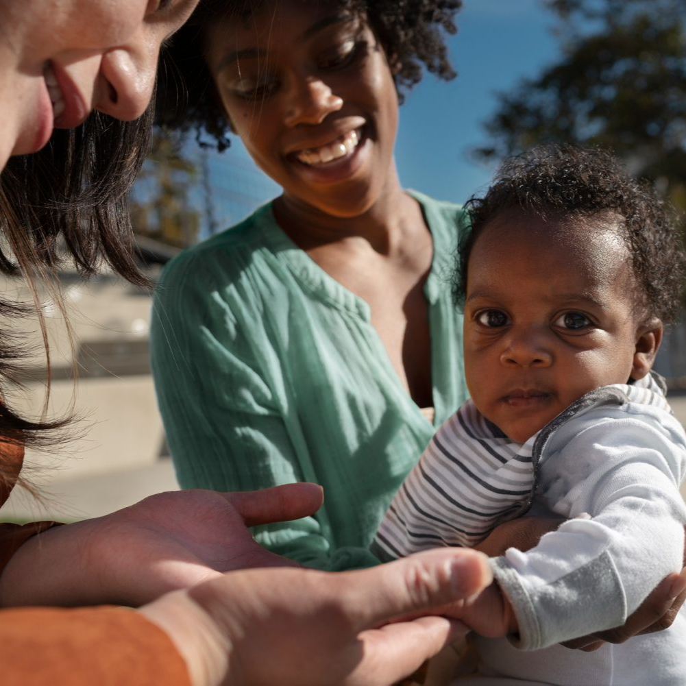 Adult holding a baby outdoors while two others smile nearby in sunlight