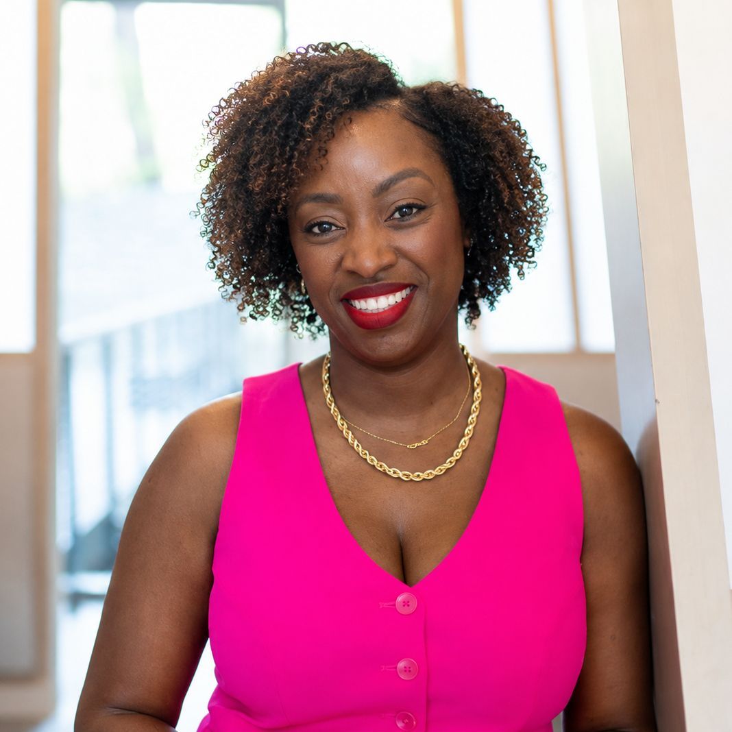 Smiling woman in a bright pink sleeveless top and gold necklace, standing indoors by a window.