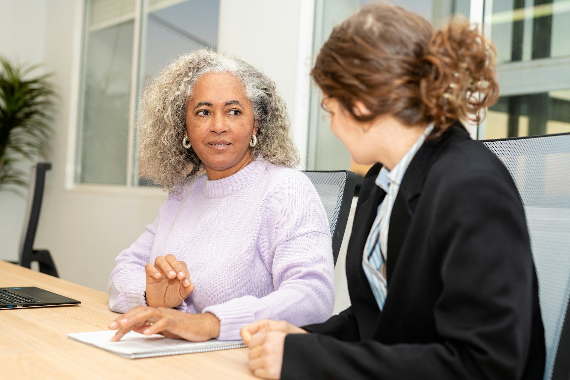 Two colleagues in an office meeting, talking across a desk with papers and a computer nearby.