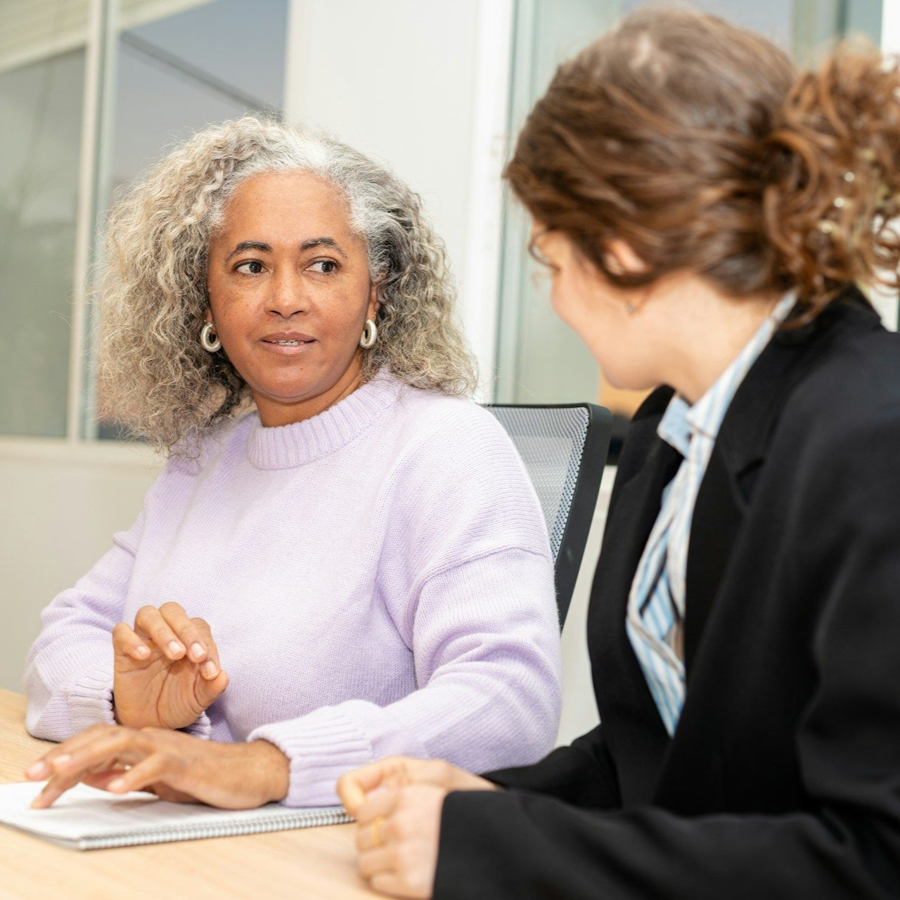 Two coworkers in discussion at a desk, one holding a notebook and the other listening attentively.