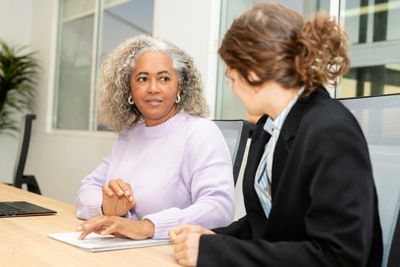Two coworkers in a meeting at a desk, one speaking while the other listens attentively