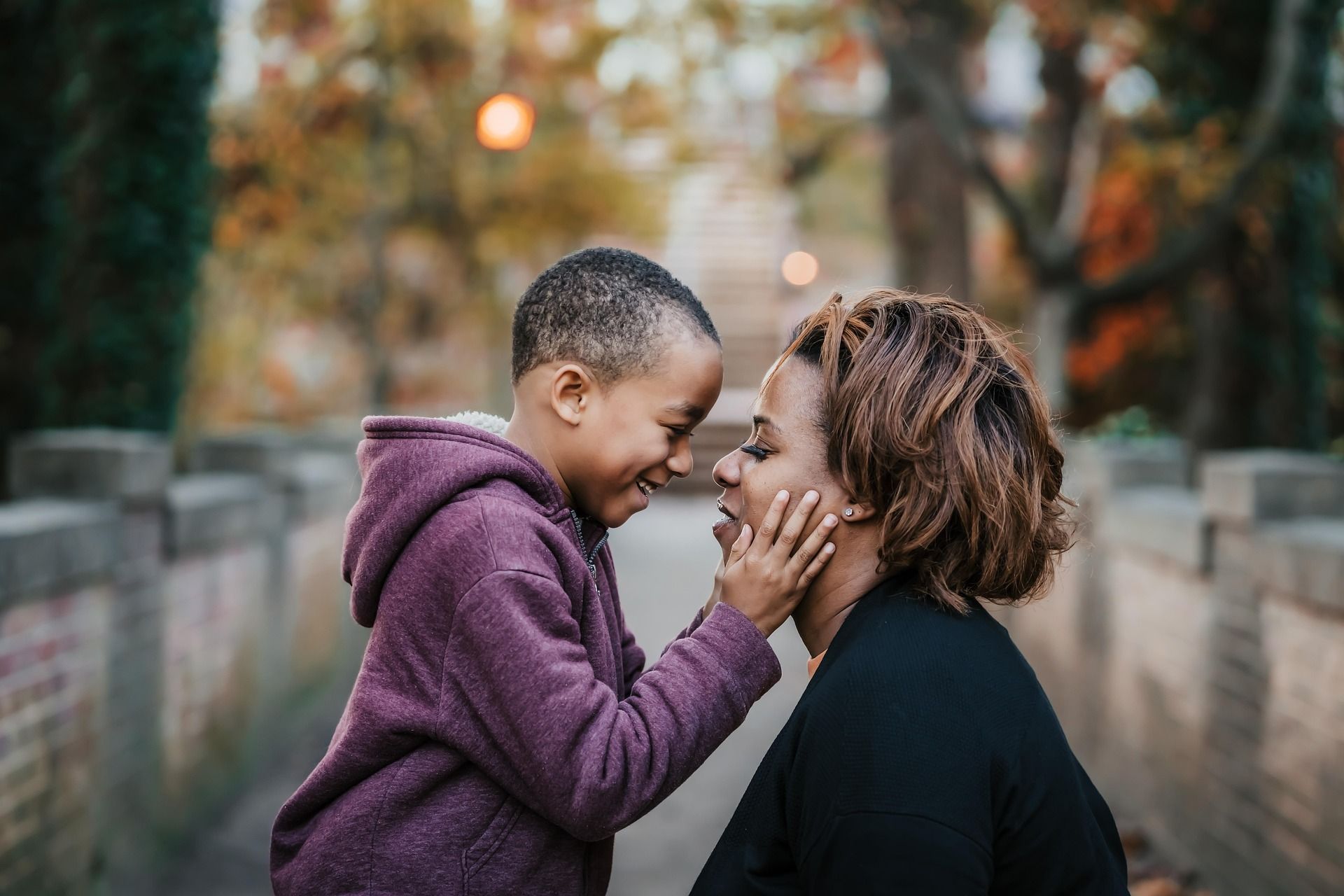 Two people smiling face to face on a tree-lined path, one gently touching the other's cheek.