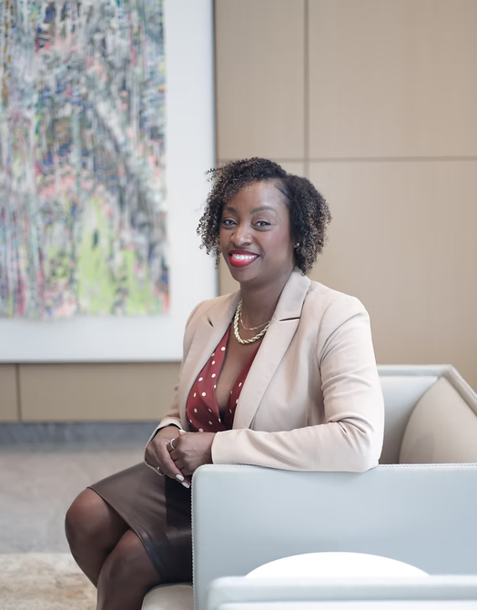 Woman seated on a white sofa in an office lobby, wearing a beige blazer and maroon top, smiling at the camera