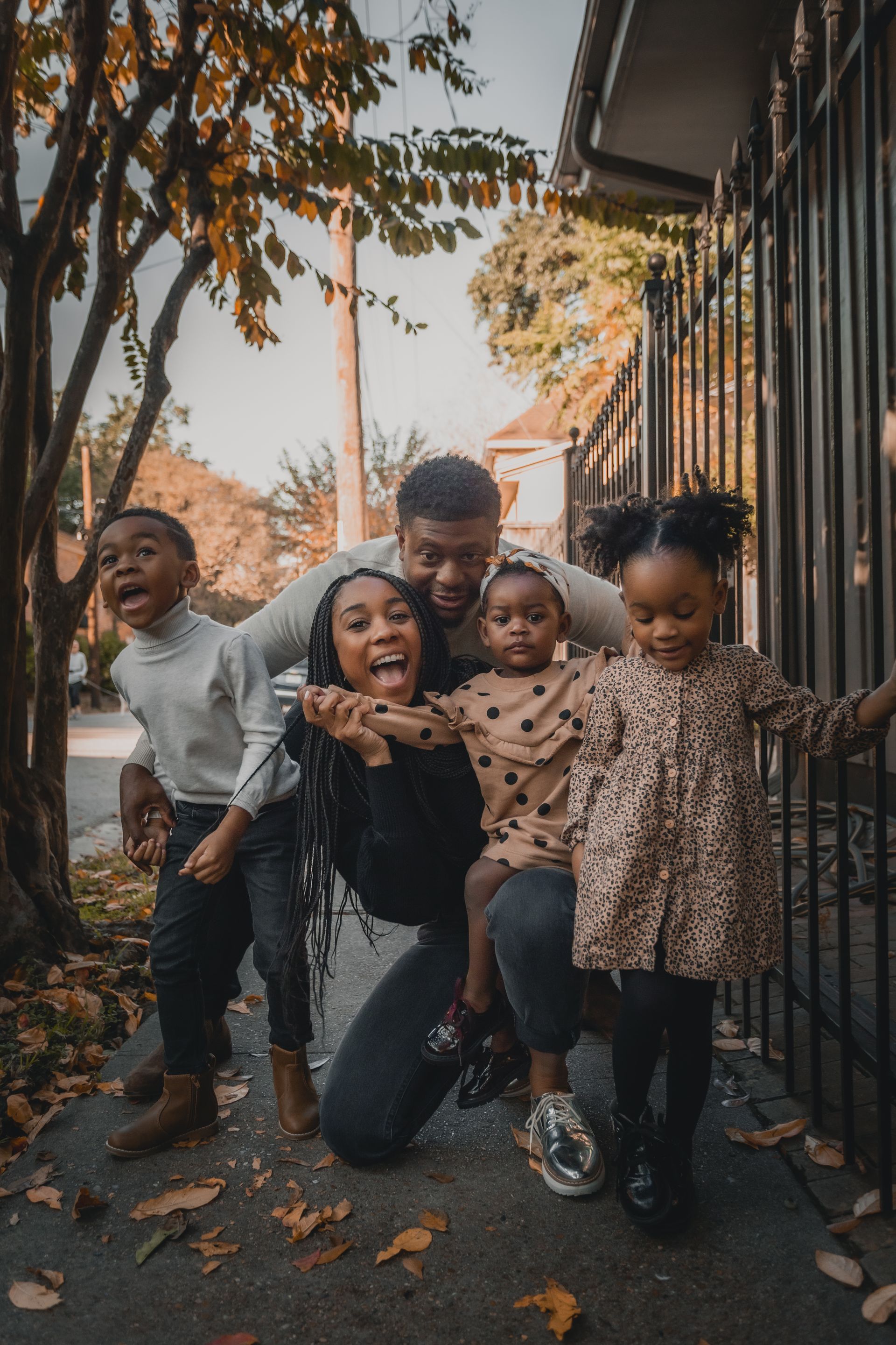 Four children smiling and posing on a leaf-strewn sidewalk beside a metal fence at sunset