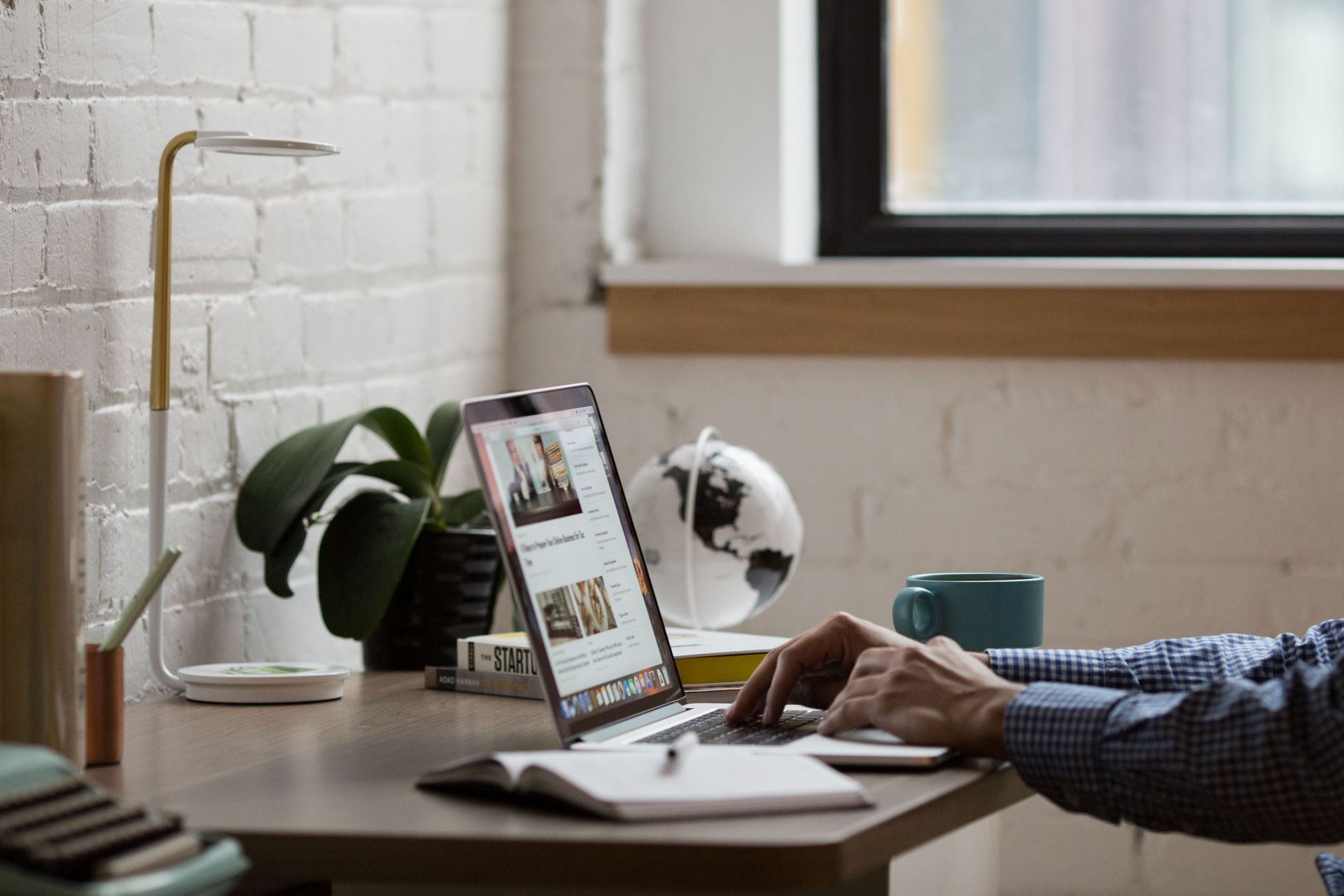Person typing on a laptop at a desk with plants and a globe by a window