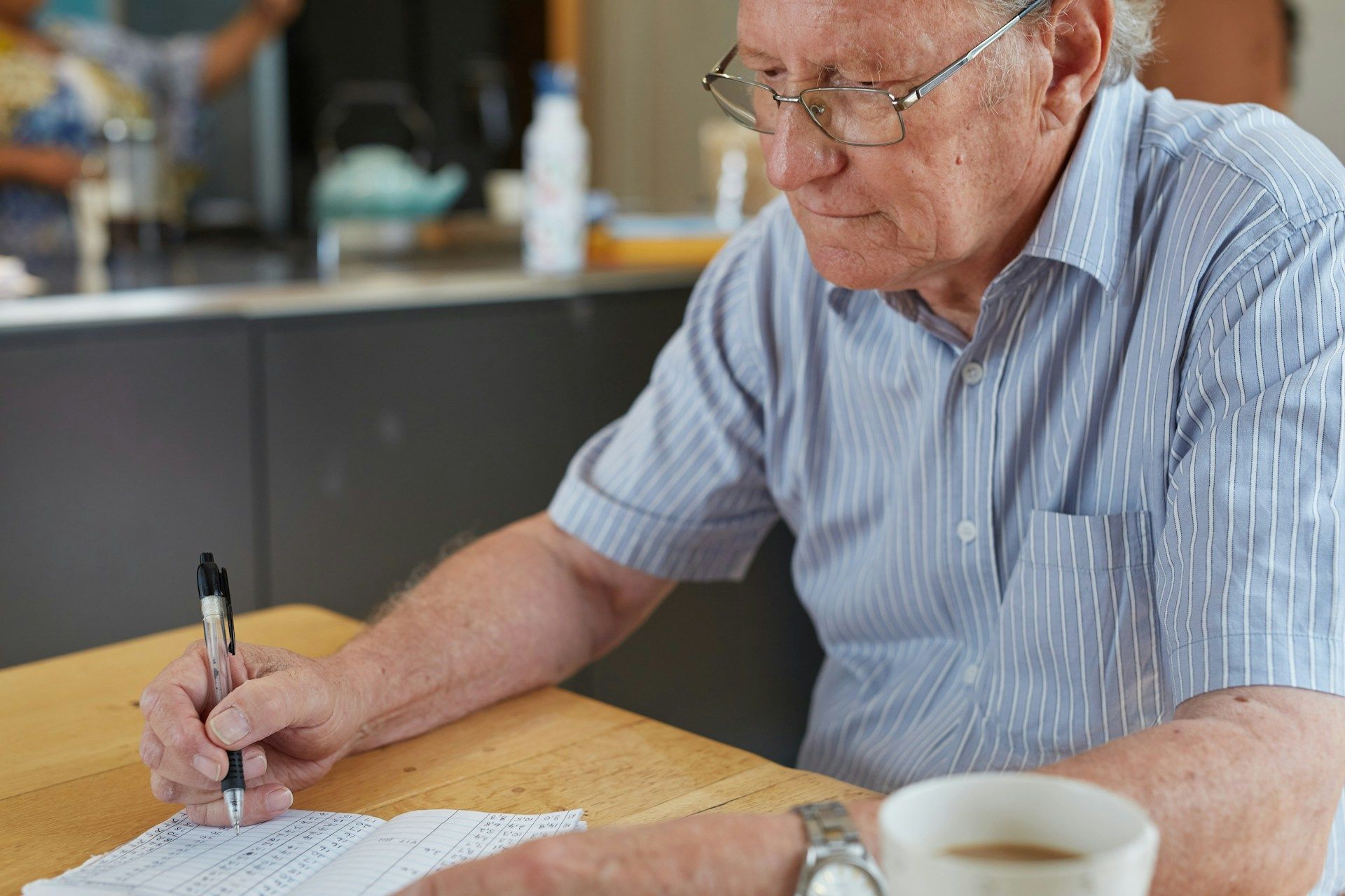 Man in blue shirt writing at a wooden table beside a coffee cup