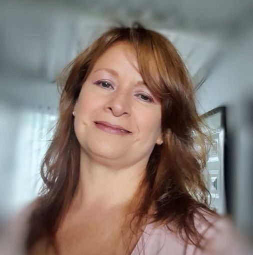 Una mujer con cabello largo color castaño rojizo sonríe, vistiendo una blusa rosa.