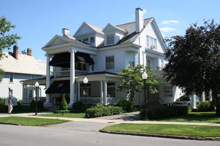 Two-story white house with covered porch and black awnings, set on a green lawn with trees, street lamps, and a sidewalk.
