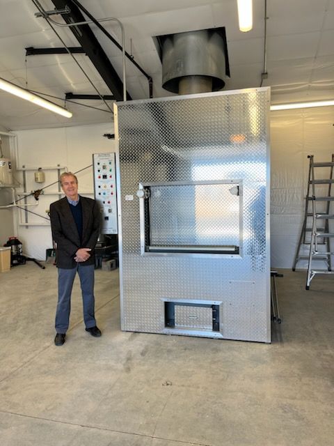 Man standing next to a large, metal industrial oven in a workshop setting.