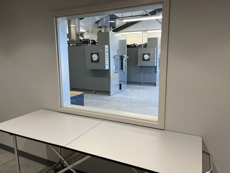 Window view of a laboratory with gray machinery, looking into a testing chamber. White table in foreground.