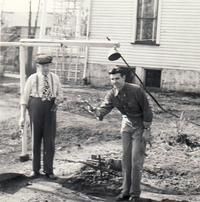 Two men outside, one holding a tool near a hole. A building is in the background.
