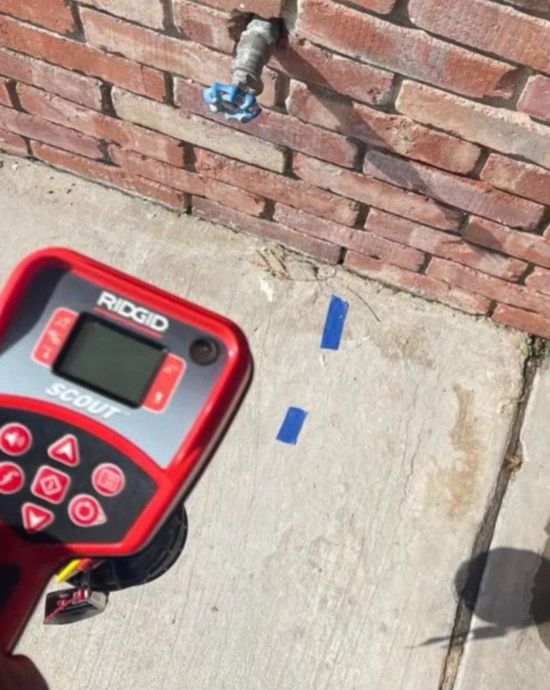 Handheld RIDGID detector aimed at concrete beside a brick wall, marking two blue tape spots on the sidewalk