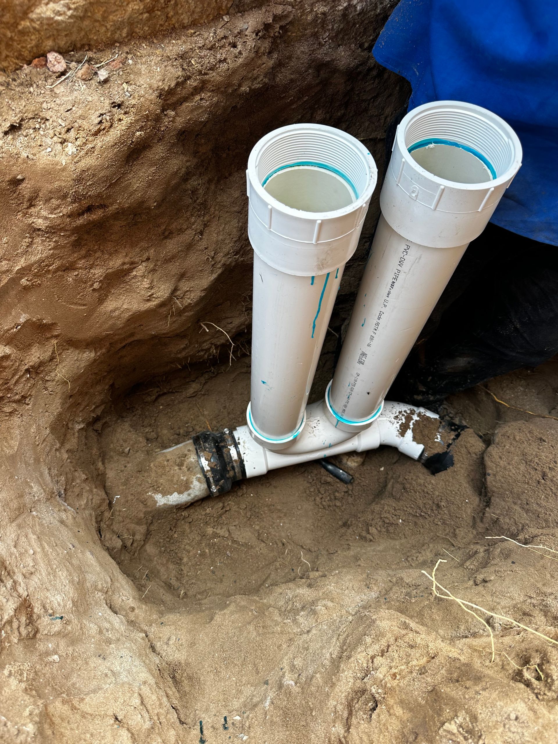 Two white PVC pipes installed in a rocky dirt trench, connected to a horizontal pipe below.