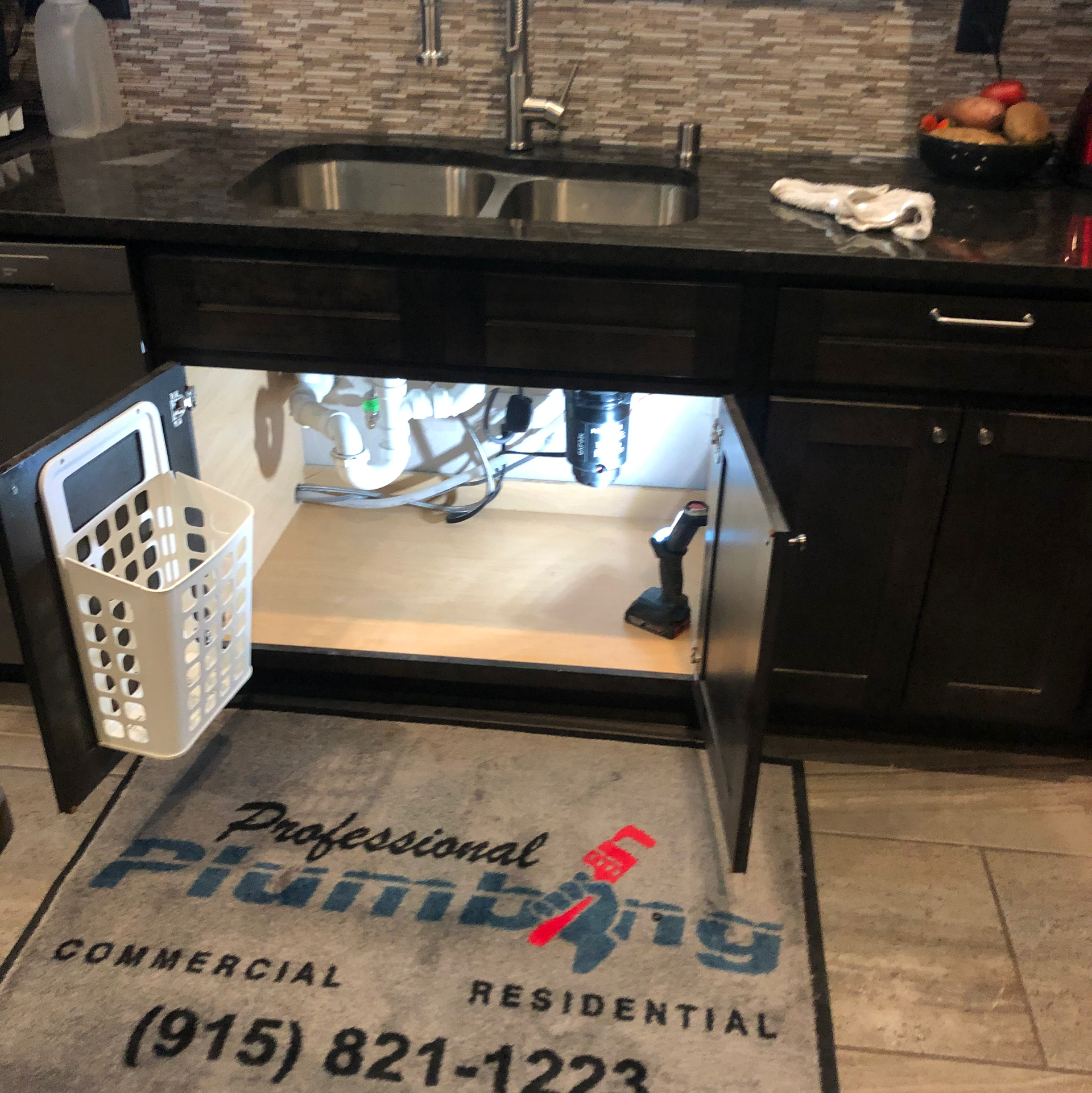 Under-sink kitchen area with sink, exposed plumbing, lighted cabinet interior, and floor mat below.
