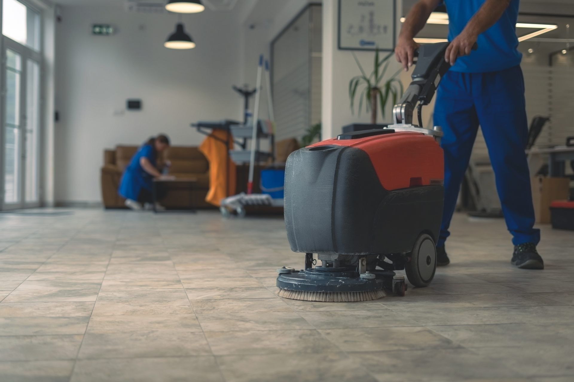 Worker using a floor scrubber in a bright indoor hallway, with another person cleaning in the background