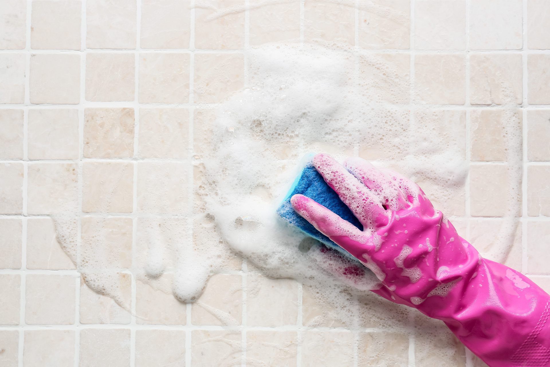 Pink-gloved hand scrubbing a tiled wall with a blue sponge, covered in soap suds