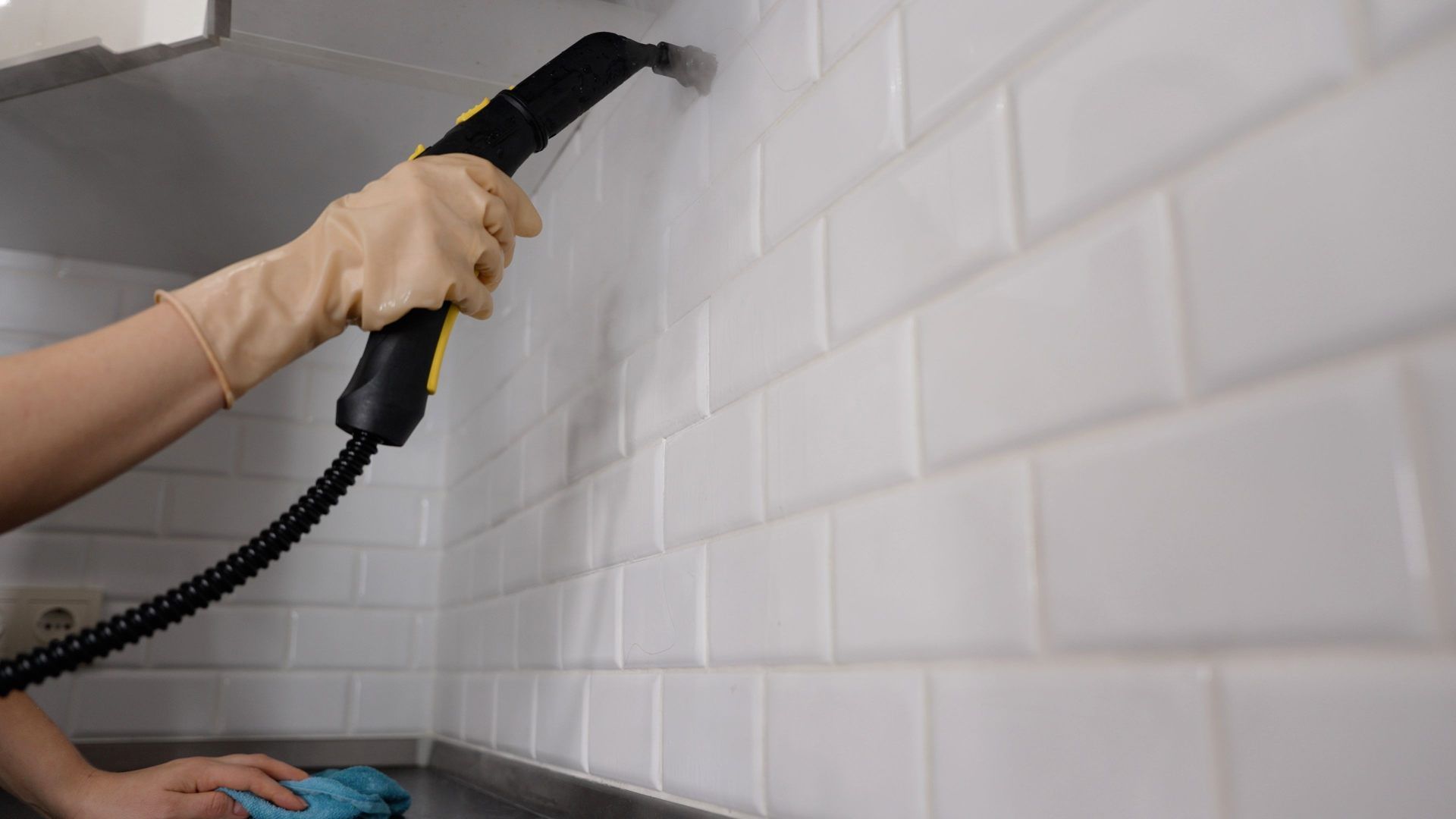 Hand using a steam cleaner to clean a white tiled wall and countertop