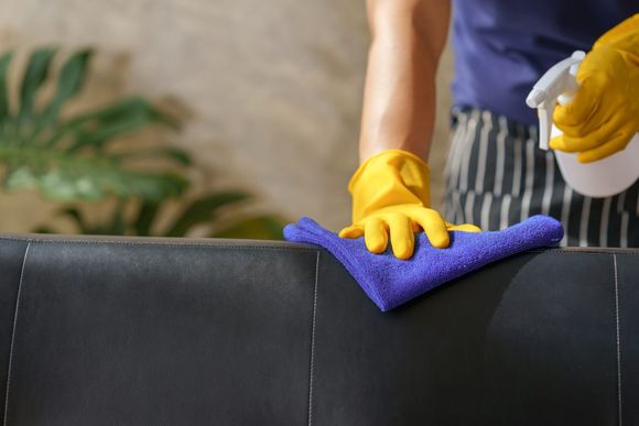 Person wiping a black sofa with a purple cloth while wearing yellow gloves