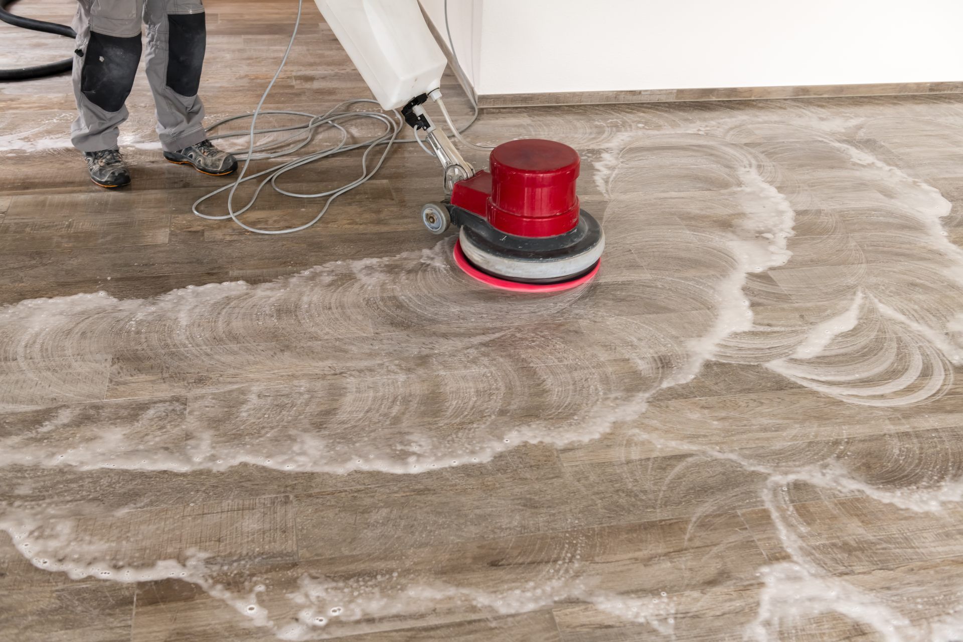 Red floor buffer polishing a dusty concrete floor with swirled dust patterns and a worker’s boots nearby
