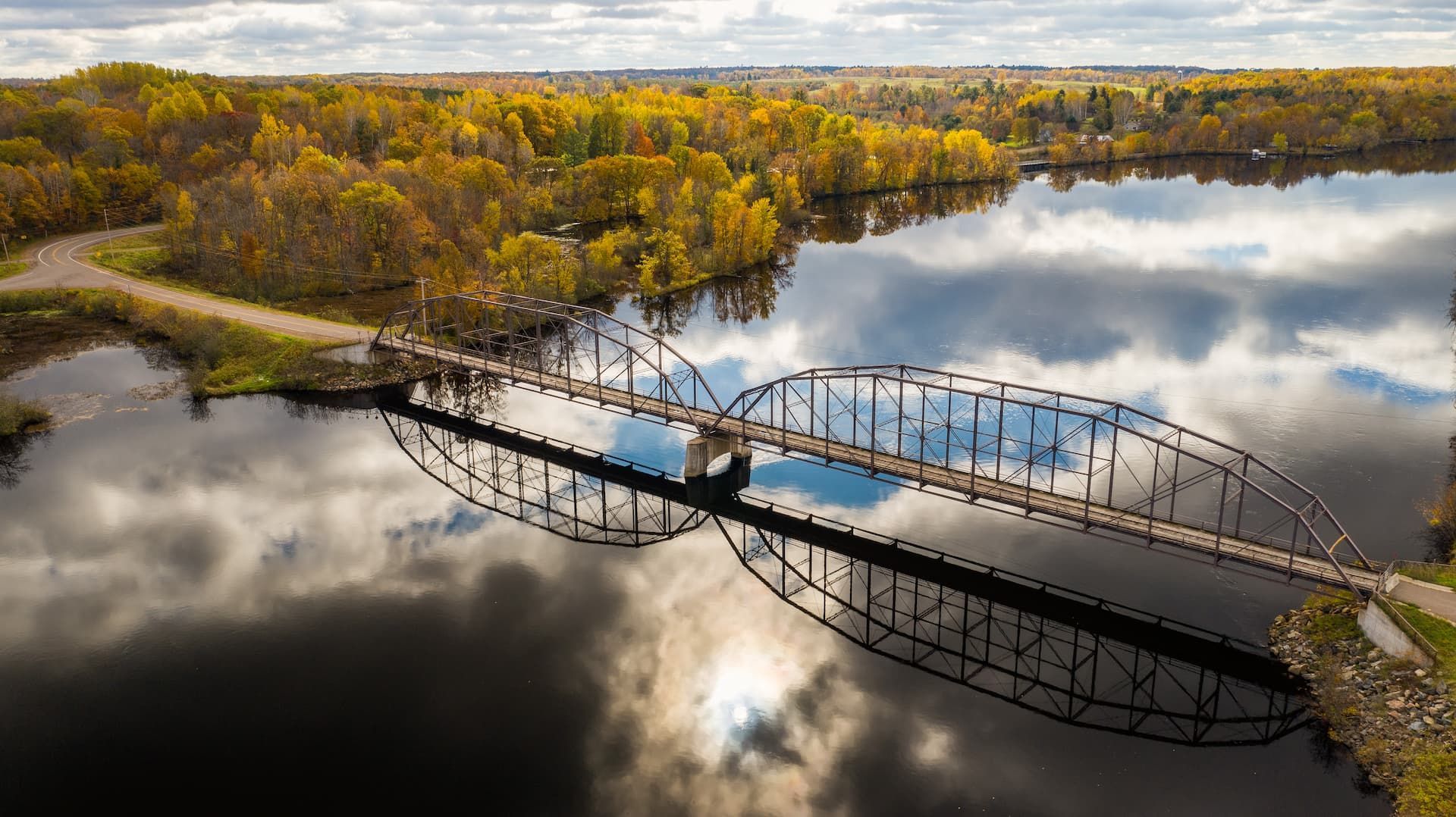 Bridge spanning a dark river reflects cloudy sky. Autumn trees line the banks.