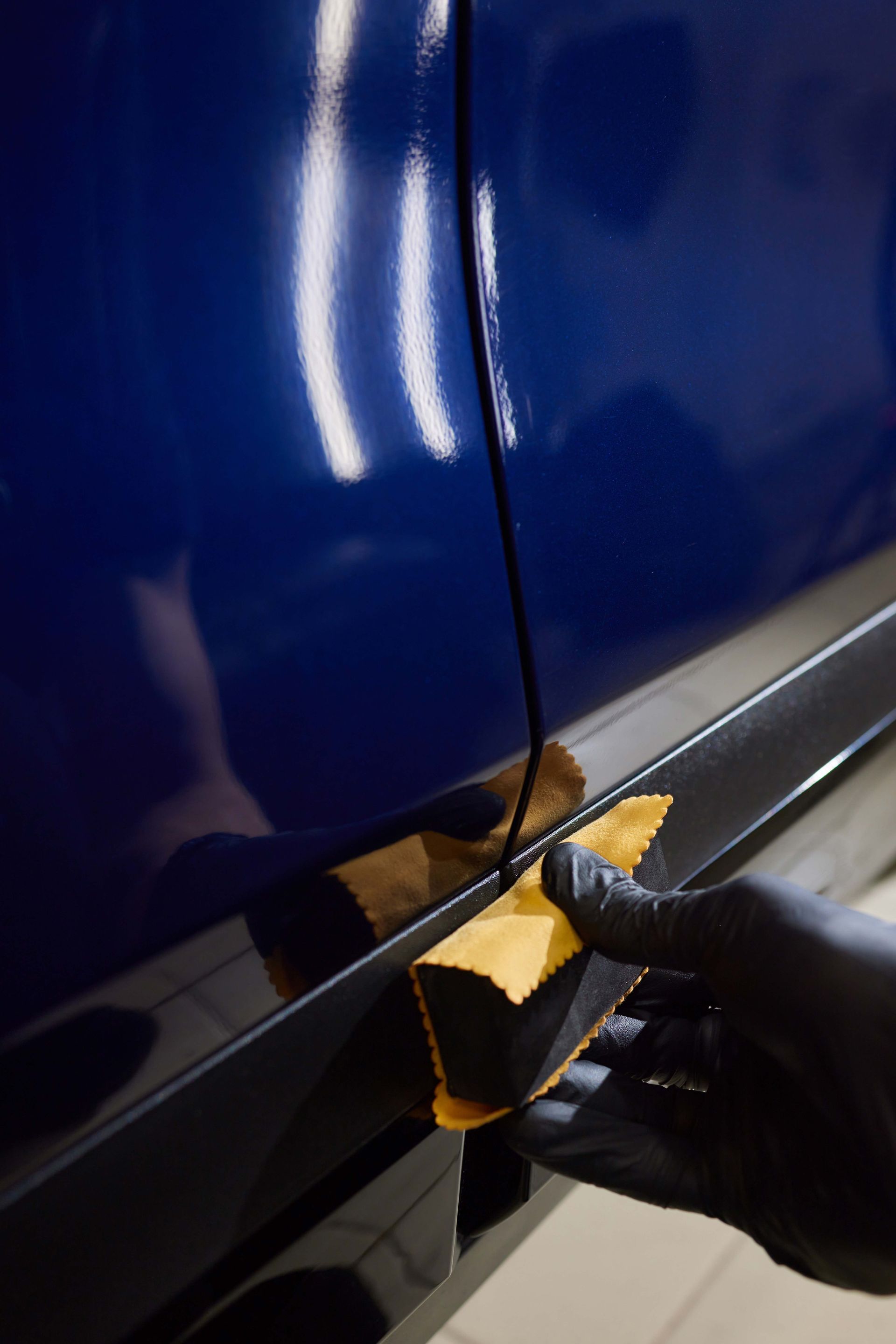 Gloved hand applies coating to black car trim with a yellow applicator; blue car body visible.