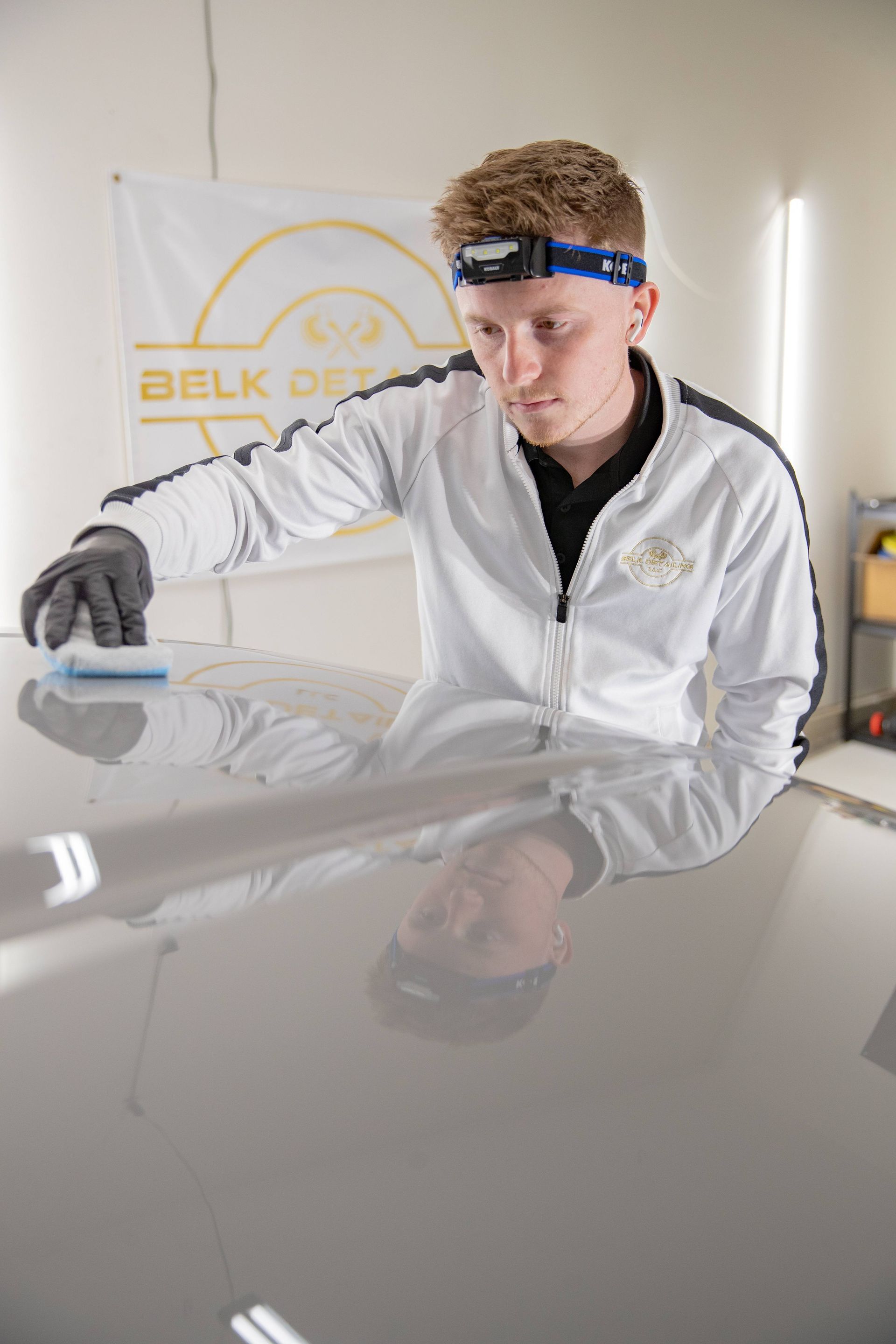Technician polishing a white car hood in a workshop, wearing gloves and a headlamp.