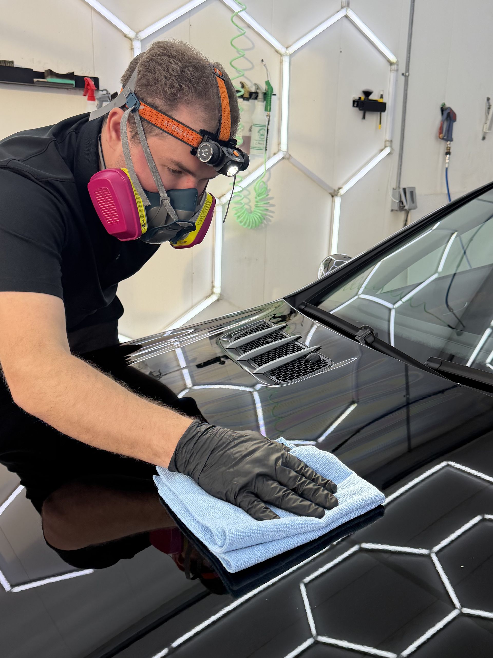 Detailing a black car hood with a cloth in a garage, wearing a respirator and gloves