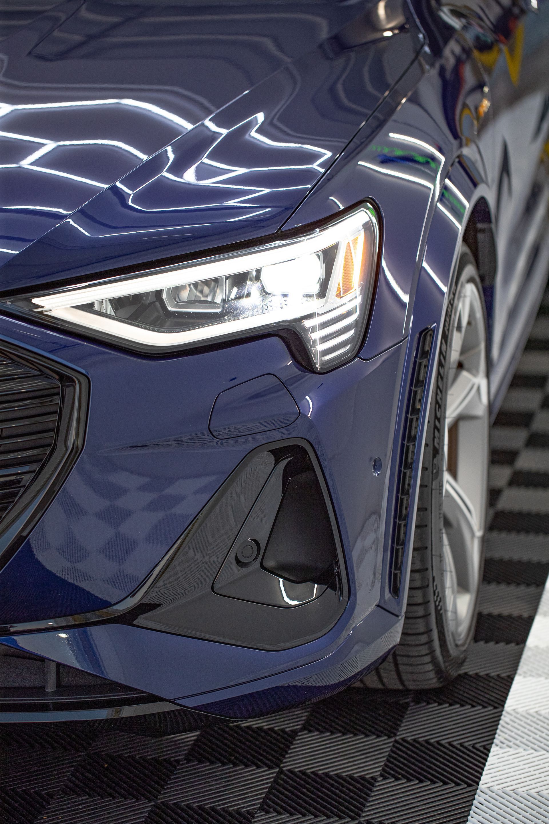 Blue Audi electric car front end with headlight, wheel, and black grill on a checkered floor.