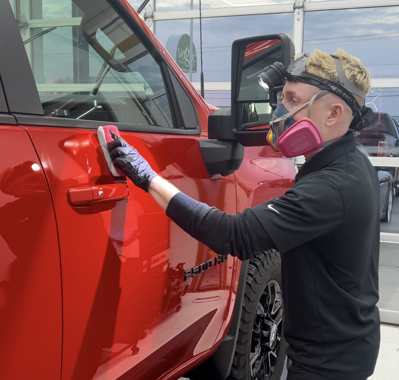 Man applying detailing product to black car hood in well-lit garage, using a microfiber cloth.
