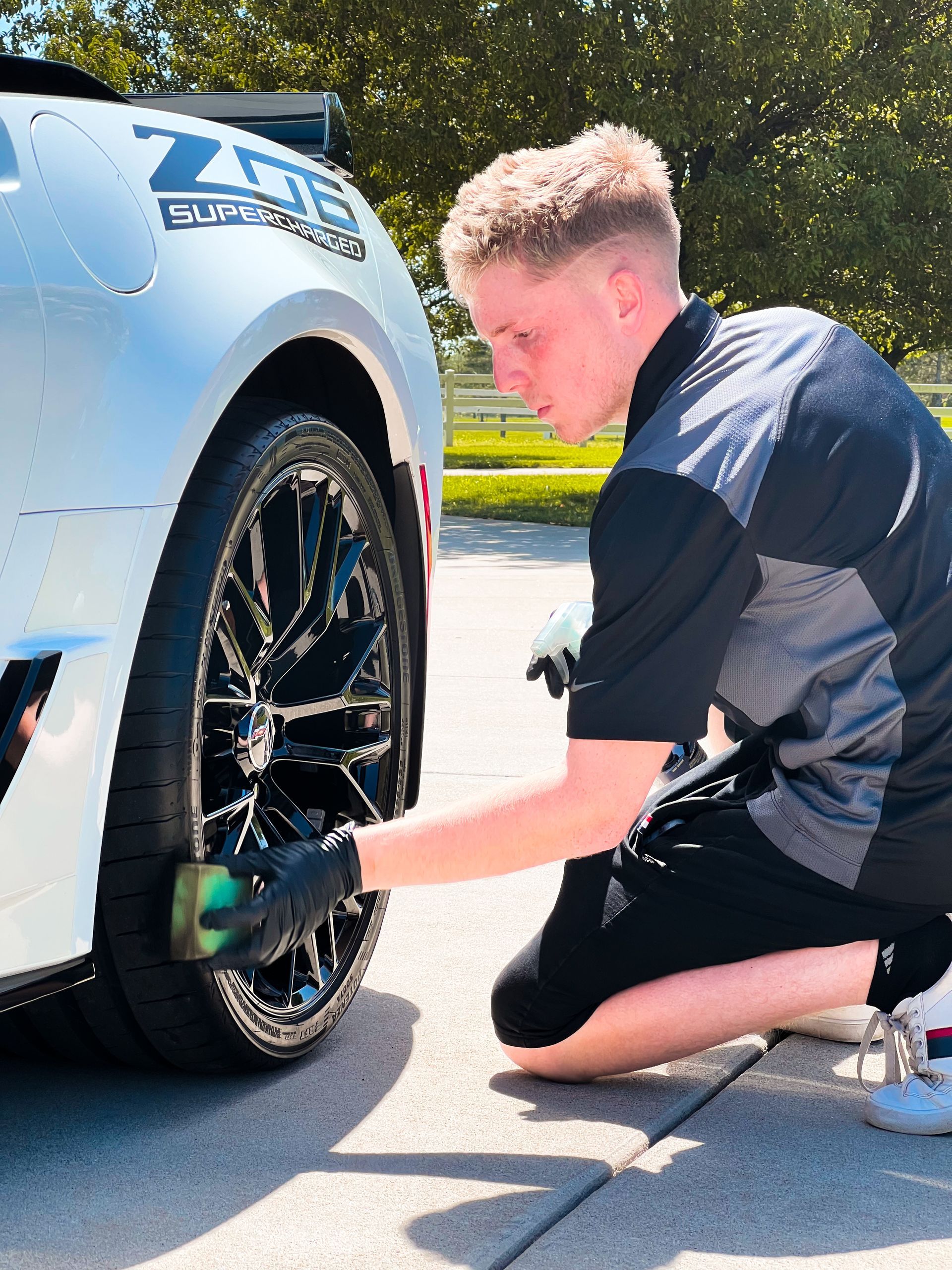 Person kneeling beside a silver car, wiping the black wheel with a cloth in bright sunlight.