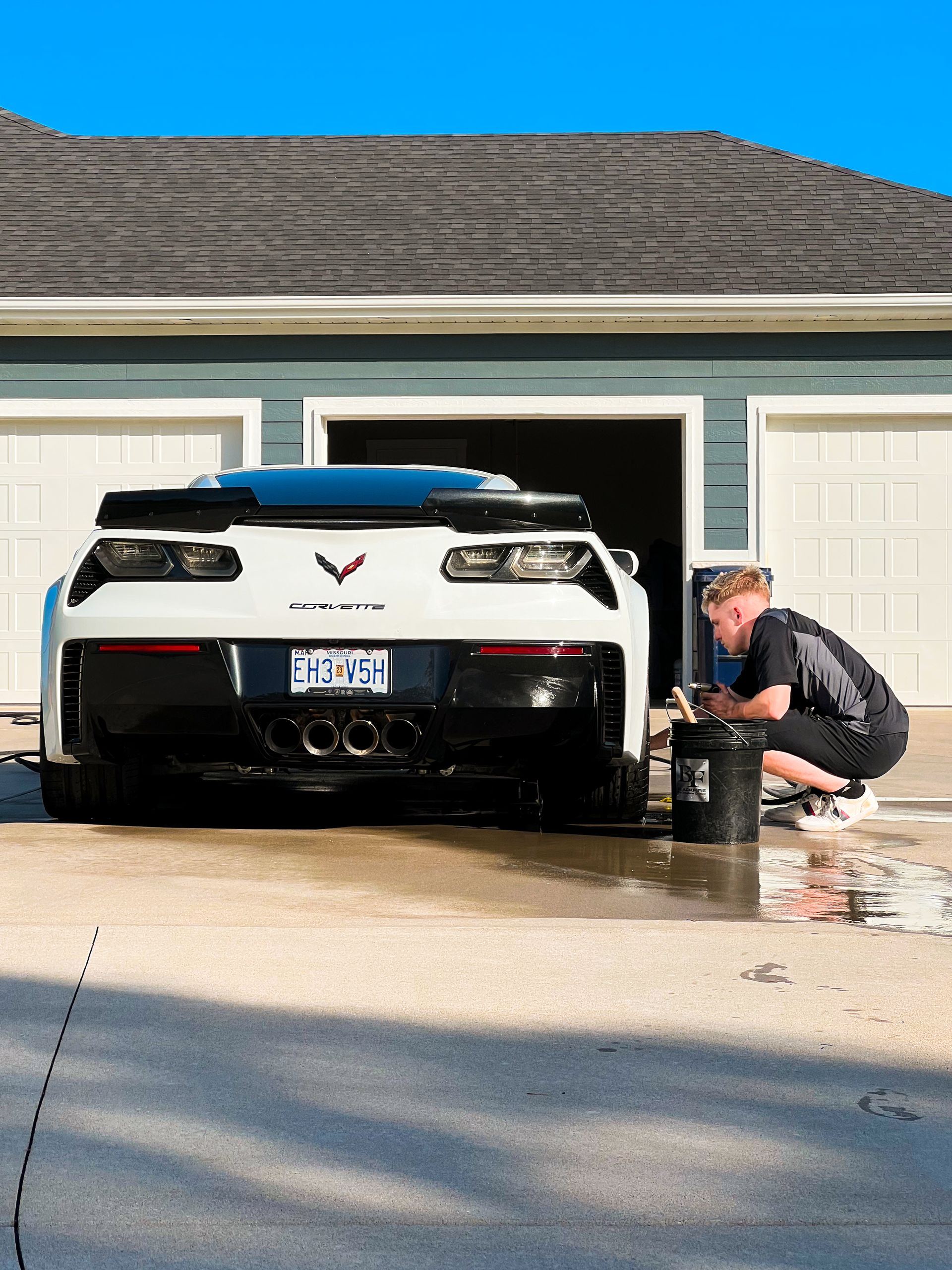 White sports car in driveway with a person washing it outside a garage.